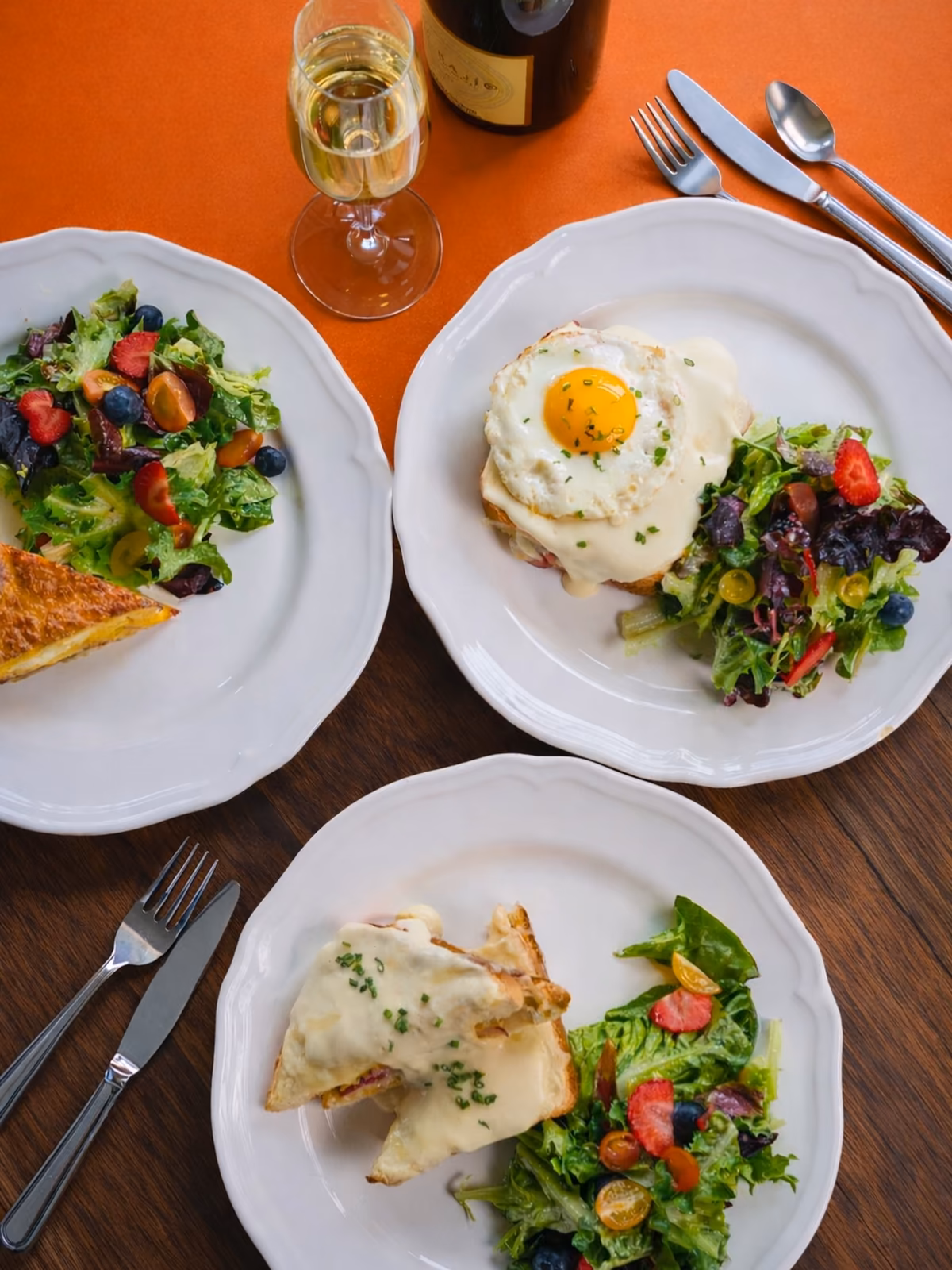 Quiche, croque madame, and croque monsieur with salads and sparkling wine on an orange table